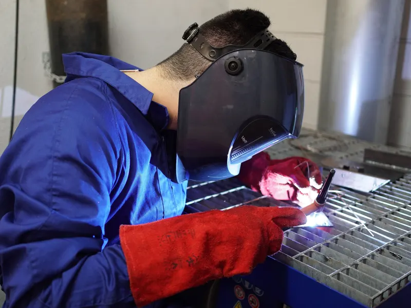 Soudeur équipé d'un masque, d'une combinaison et de gants de soudeur en cuir rouge, penché sur une grille